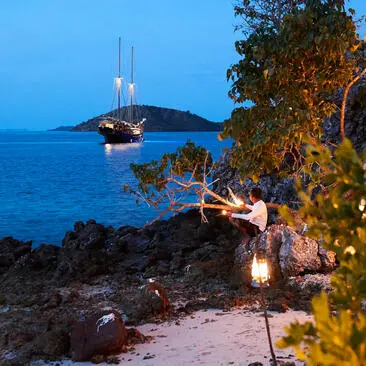 Amandira's waterfront at dusk, with illuminated lanterns along the rocky shore and a moored sailing vessel on calm blue waters.