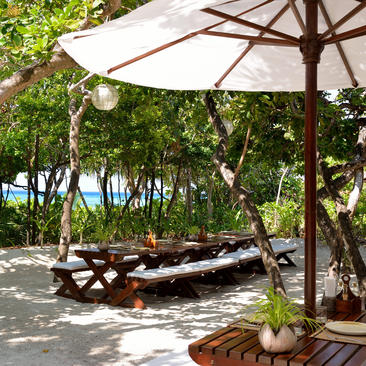 Shaded dining area at Amanpulo with wooden lounger beneath white parasol, overlooking tropical gardens and ocean.