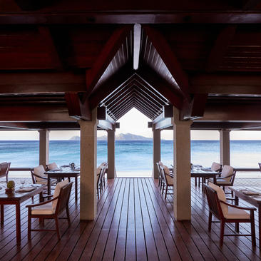 Amanpulo's Beach Club dining venue with wooden beams, set tables, and ocean views through floor-to-ceiling windows.