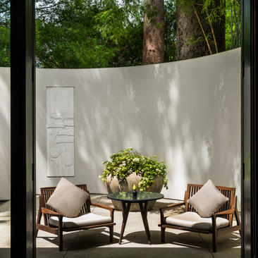 Shaded seating area at Amansara with two wooden chairs, cushions, and potted plant against white wall.