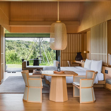 Amanemu suite with wooden furniture, pendant lighting, and open views to landscaped gardens through floor-to-ceiling windows.