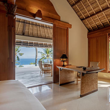 Ocean Suite bedroom at Amankila resort with open doors to ocean views, wooden furnishings and terracotta roof details.