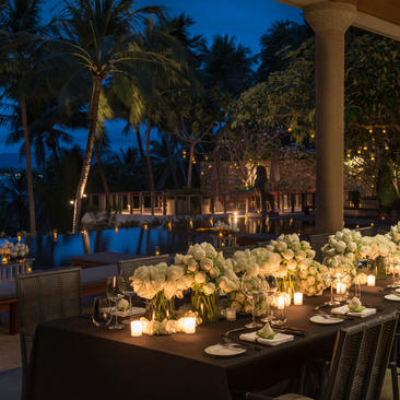 安缦璞瑞度假村的事件用餐区夜景，长餐桌两侧点缀着烛光，棕榈树和建筑柱子映衬其中。
