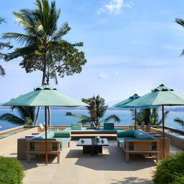 安缦璞瑞度假村的海滨露台，配有木制座椅、遮阳伞和棕榈树，俯瞰清澈海水。