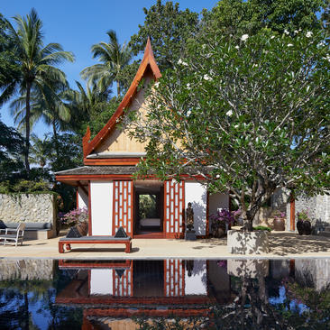 Amanpuri resort's traditional Thai pavilion reflected in still water, framed by palm trees and lush foliage.