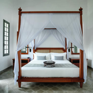 Khan Suite principal bedroom at Amantaka, featuring a four-poster bed with white drapes and wooden bedside tables.