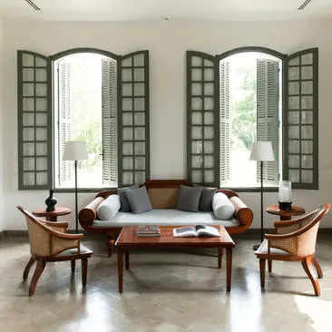 Sitting room at Amantaka with symmetrical arched windows, mid-century wooden chairs, and cushioned sofa beneath gridded shutters.