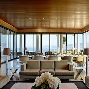 Villa living area at Amanzoe resort with floor-to-ceiling windows overlooking the Aegean Sea, neutral furnishings and wooden ceiling.