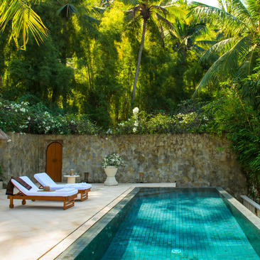 Sunlit plunge pool at Amankila with loungers beside a stone wall, surrounded by lush tropical vegetation.