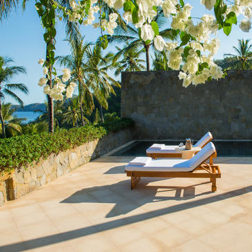 Wooden daybed with cushions on a sun-drenched terrace at Amankila, overlooking tropical gardens and ocean views.