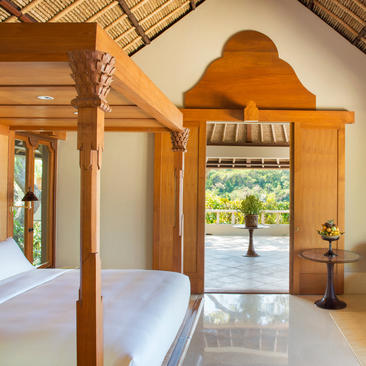 Wooden four-poster bed in a light-filled bedroom at Amankila, with open doors framing a view of tropical gardens beyond.