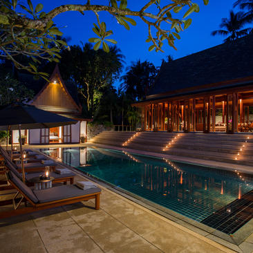 Amanpuri resort at dusk, illuminated pavilions and pool reflecting warm light beneath a twilight sky.