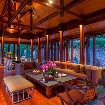 Amanpuri resort dining pavilion with warm wooden beams, floor-to-ceiling windows overlooking evening gardens, and vibrant fuchsia floral arrangements.