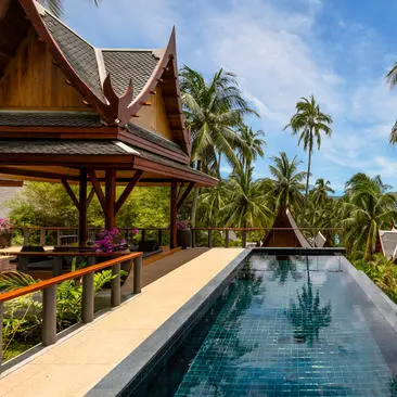 Amanpuri's infinity pool extends towards tropical gardens and traditional Thai architecture beneath clear skies.
