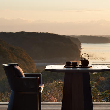 Dining table with chair overlooking forested coastline at Amanemu at sunset.