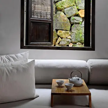 Amanfayun villa interior with grey sofa, white pillows, and wooden table framed by window overlooking stone courtyard.