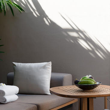 Amanwella suite detail showing cushioned seating, wooden side table with green vessel, and palm shadow on grey wall.