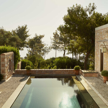 Amanruya pavilion with rectangular plunge pool overlooking partial sea view, framed by stone walls and pine trees.