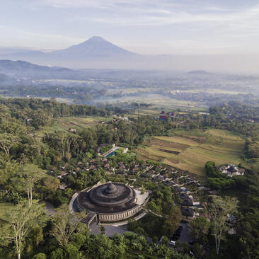 Aerial view of Amanjiwo's circular architecture surrounded by lush vegetation with Mount Merapi in the distance, Indonesia.