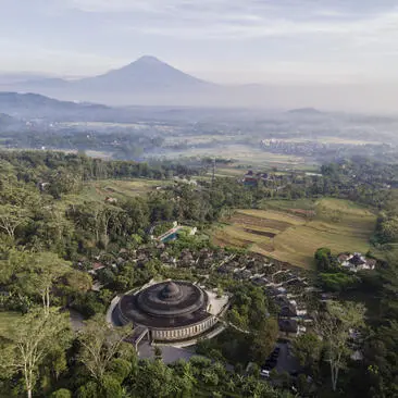 Aerial view of Amanjiwo's circular architecture surrounded by lush vegetation with Mount Merapi in the distance, Indonesia.