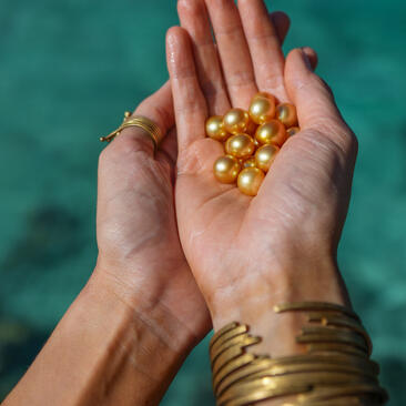 Two hands holding golden pearls above turquoise water at Amanpulo resort.