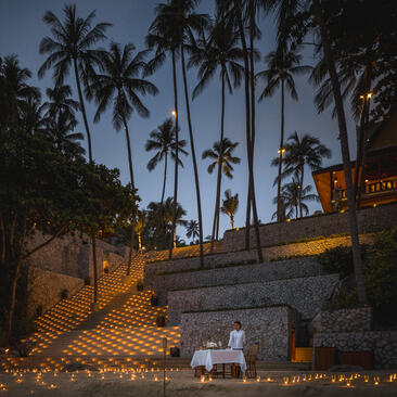 Amanpuri resort at dusk, with tall palm trees illuminated against darkening sky and warm lights glowing from stone structures below.