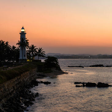 Galle Lighthouse stands on a rocky headland at sunset, its beacon illuminated against a soft peachy sky, with palm trees and calm waters below at Amangalla.