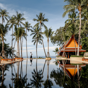 Amanpuri's infinity pool reflects golden pavilions and palm trees under clear skies.