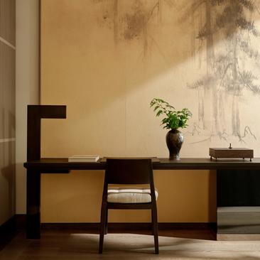 Minimalist workspace at Aman New York with wooden desk, chair, and potted plant against pale wall.