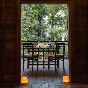 Dining table and chairs on a terrace at Amansara, framed by open wooden doors with lanterns on either side, overlooking lush greenery.