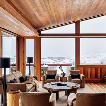 Wooden interior at Amangani with expansive windows overlooking snow-covered landscape and seated guests by fireplace.
