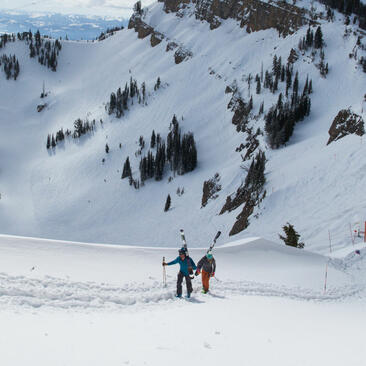 Two skiers descend a snow-covered mountain slope at Amangani, surrounded by evergreen trees and alpine terrain.