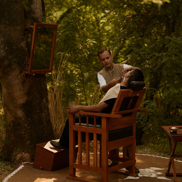 A guest sits alone in a wooden chair beneath towering trees at Aman-i-Khas, surrounded by dappled sunlight filtering through the canopy.