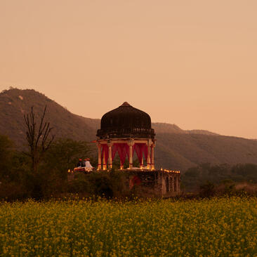 Chhatri dinner pavilion at Amanbagh, illuminated at dusk in a mustard field with mountains beyond.