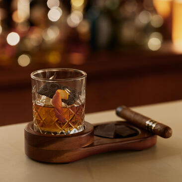 Glass of amber spirit with ice on wooden coaster at Amanera's dining venue, beside cigars.