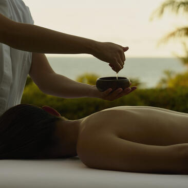 Spa therapist pouring oil onto a guest's shoulder at Amanera, with sea views beyond.