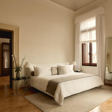 Bedroom with frescoed ceiling at Aman Venice hotel, featuring a bed with neutral linens and large windows overlooking the city.