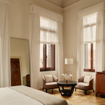 Dormitorio con techo alto, cama blanca y zona de estar con sillones junto a ventanales en Aman Venice.