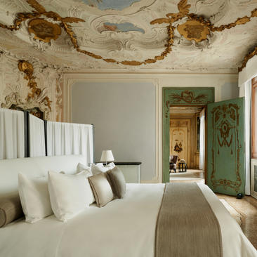 Alcova Tiepolo Suite principal bedroom at Aman Venice, with ornate ceiling and neutral décor.