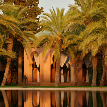 Moroccan resort gazebo framed by palm trees and their reflections in still water at dusk, Amanjena.