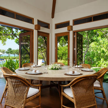 Dining room at Amanpulo resort with round table, rattan chairs, and tropical views through large windows.