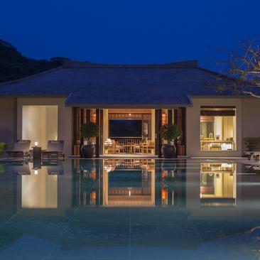 Amanoi pavilion illuminated at dusk, reflected in still water, with mountains beyond.
