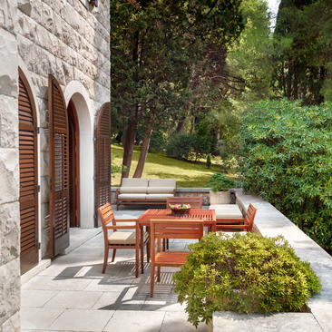 Wooden dining table and chairs on a stone terrace at Aman Sveti Stefan, surrounded by lush greenery and arched stone architecture.