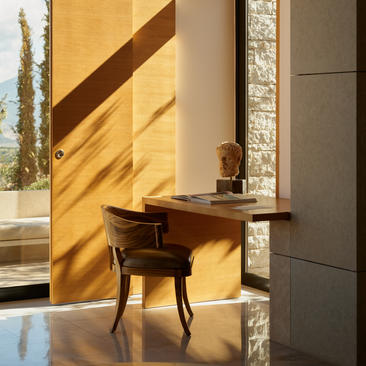 安缦卓熠度假村的泳池馆内部，木制书桌沐浴在阳光下，旁边是石墙和落地窗。