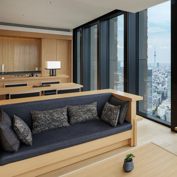 Panorama Suite lounge at Aman Tokyo with floor-to-ceiling windows overlooking the city skyline, featuring a built-in wooden bench with dark cushions and a desk area.