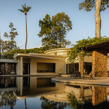 Amansara's exterior reflected in still water, with palm trees and stone architecture framed by clear sky.