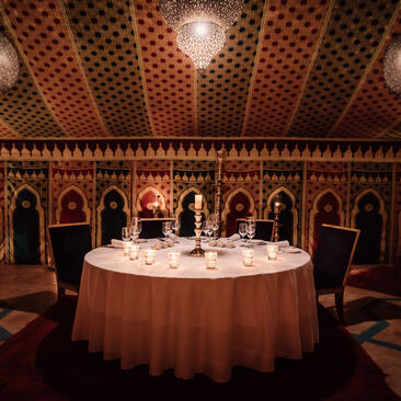 Caïdal tent dining room at Amanjena resort with round table set for dinner, ornate carved wooden walls and soft ambient lighting.