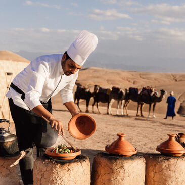 Man in white clothing pouring water into clay vessels in the Moroccan desert at Amanjena, with camels and handlers in the background.