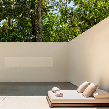 Wooden daybed with cushions on a shaded terrace at Amansara, surrounded by lush greenery.