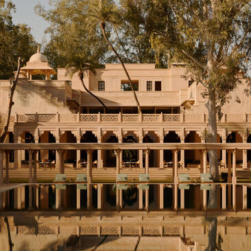 Amanbagh's main building reflected in the pool, with arched colonnades and warm stone architecture framed by trees.